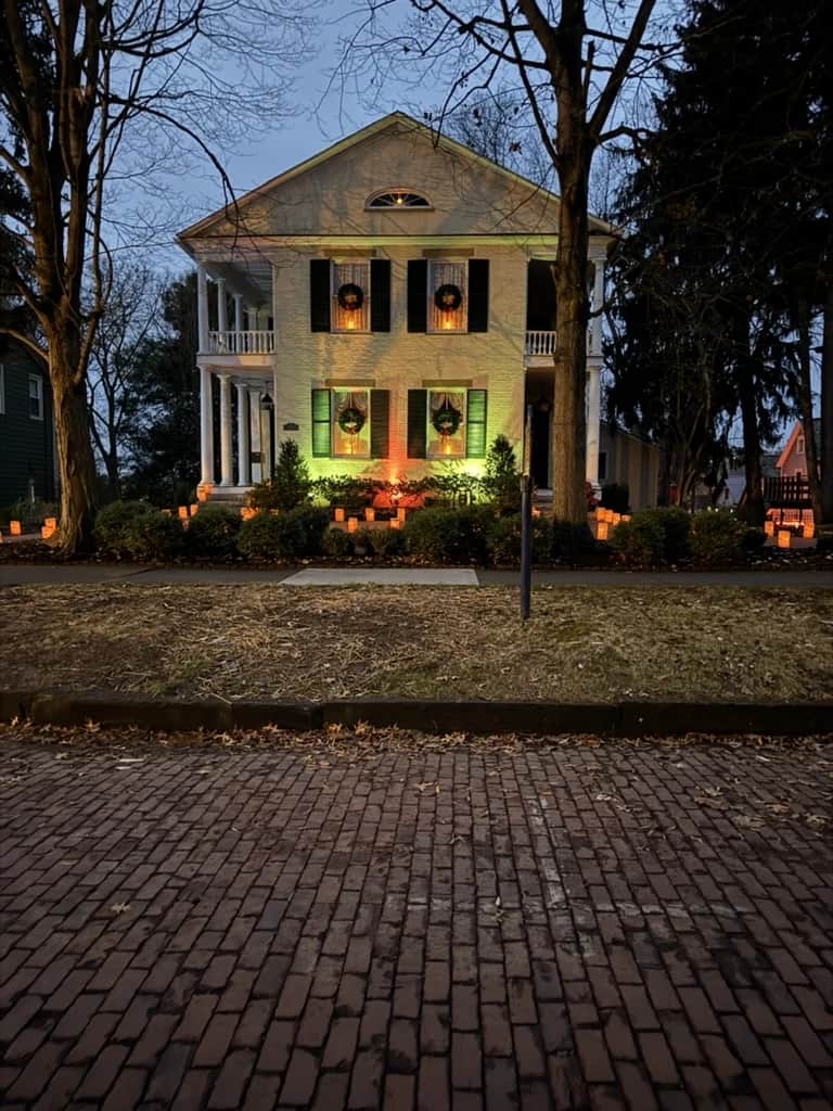 The house seen from the brick street at winter dusk, surrounded by luminaria